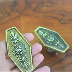 Op de afbeelding: Twee sierlijke messing ladegrepen met een bloemmotief. De grepen zijn zeshoekig en hebben een reliëf.