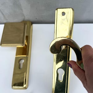 Puede incluir: Dos manillas de puerta doradas, una con una placa cuadrada y la otra con una manija curva. Ambas manillas tienen mirillas.