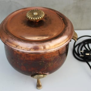 Könnte beinhalten: Ein antiker elektrischer Speisenwärmer aus Kupfer mit einem Messingknopf auf dem Deckel und drei Messingfüßen. Der Wärmer hat ein Kabel und einen Stecker.