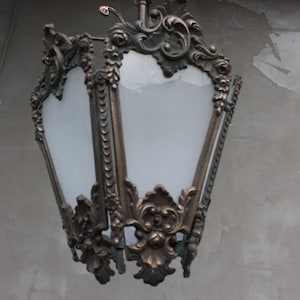 Puede incluir: Lámpara colgante de color bronce ornamentada con un panel de vidrio esmerilado. La lámpara tiene un diseño decorativo y vintage con detalles florales y de volutas. Los paneles de vidrio son blancos y el marco de metal tiene un aspecto envejecido.