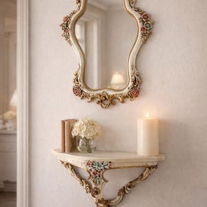 May include: Ornate, white and gold wall mirror with floral accents, hanging above a matching shelf. The shelf holds a vase of flowers, books, and a lit candle. The decor has a vintage, romantic aesthetic.