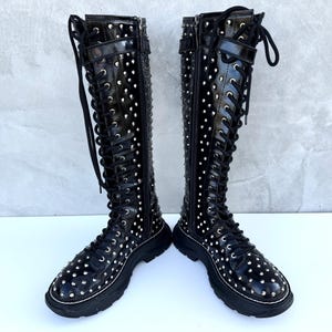 Może przedstawiać: Para czarnych skórzanych kozaków za kolano z srebrnymi ćwiekami i sznurowaniem.