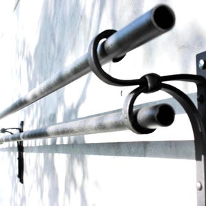 Puede incluir: Un juego de barras de cortina dobles de hierro forjado contra una pared blanca. Las barras son de color plateado texturizado, con soportes circulares y curvos decorativos. El diseño es minimalista e industrial.