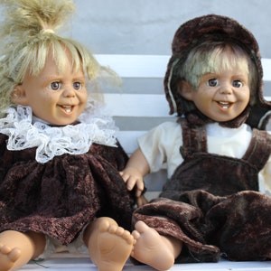 May include: Two dolls sitting on a white bench. The doll on the left is wearing a brown dress with a white lace collar and has blonde hair. The doll on the right is wearing a brown and white outfit with a brown hat and has blonde hair.