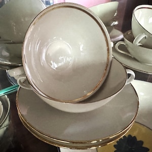 Puede incluir: Juego de cuencos de sopa y platillos vintage de color crema con ribete dorado. Los cuencos tienen dos asas y están apilados. Se ven otras tazas y platillos, lo que sugiere un juego de vajilla completo.