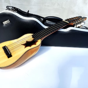 Puede incluir: Un charango de color claro, un pequeño instrumento de cuerda andino, descansa frente a un estuche negro. El charango tiene un cuerpo de madera con una boca y varias cuerdas. El estuche tiene un ribete plateado y la palabra "Caprice".