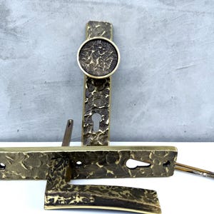 May include: A set of antique brass door hardware, including a handle, keyhole plate, and a decorative escutcheon. The pieces have a textured, hammered finish and a warm, aged patina. The set includes screws for installation.