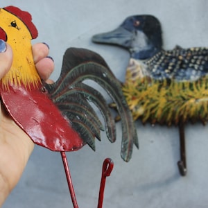 May include: Two metal wall hooks shaped like a rooster and a loon. The rooster is red, yellow, and black. The loon is blue, black, and yellow.