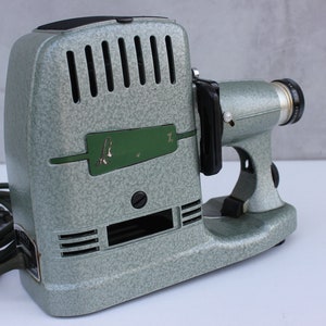 Könnte beinhalten: Ein grüner Vintage-Diaprojektor mit schwarzem Griff und Objektiv. Der Projektor hat ein Kabel und ein grünes Etikett mit dem Text "A.X."