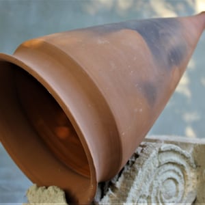 Puede incluir: Una maceta de terracota con forma de cono con una abertura ancha en la parte superior. La maceta está apoyada sobre una superficie de piedra.