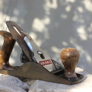 Op de afbeelding: Een vintage houten handvlak met een messing body, een houten handvat en een metalen mes. Het mes heeft een rood label met de tekst "ANANT".