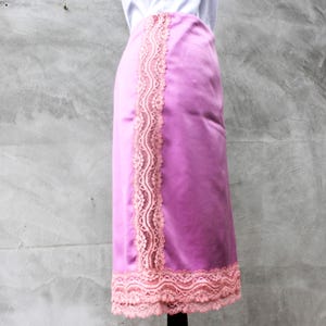 May include: A lilac satin skirt with light pink lace trim along the side and hem. The skirt is displayed on a mannequin against a grey background. The lace adds a touch of elegance.