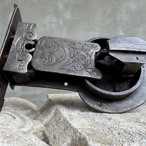 Puede incluir: Un pestillo de puerta de metal antiguo y ornamentado con intrincados grabados florales. El pestillo es de metal oscuro y envejecido, con una placa rectangular, un brazo giratorio y una carcasa circular. El diseño sugiere una estética vintage o histórica.