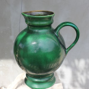 May include: A large, green ceramic pitcher with a rounded body and a wide, flared lip. The pitcher has a single handle and a slightly raised base.