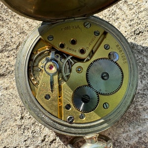 May include: An open Omega pocket watch, revealing its intricate mechanism. The interior showcases gold-coloured gears, a ruby jewel, and the markings "OMEGA SWISS 15 JEWELS." The serial number "8581265" is also visible.
