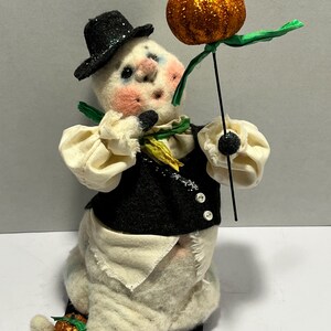 May include: A whimsical Halloween decoration featuring a ghost-like figure. The figure wears a black hat and vest over a cream-colored outfit. It holds a glittery orange pumpkin on a stick. The figure has rosy cheeks and a small pumpkin on its foot.