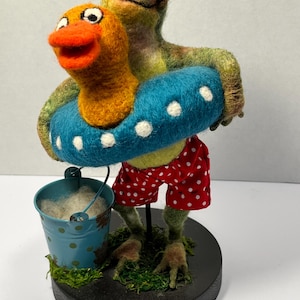 May include: A whimsical felt frog figurine wearing red polka dot shorts and holding a blue inflatable ring with a yellow duck. A small blue bucket with white contents sits nearby. The figurine is on a black base with green moss.