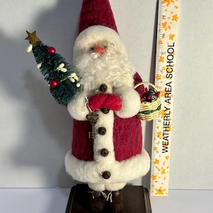 May include: A handcrafted Santa Claus figurine, approximately 6 inches tall, with a red and white felt outfit. The figure holds a small Christmas tree and a basket. A key hangs from his hand. The base is dark brown.