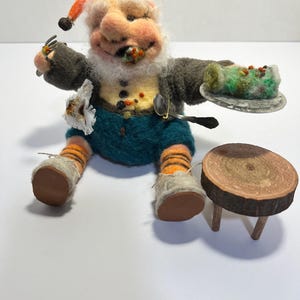 May include: A whimsical felted gnome figurine with an orange hat, gray jacket, and teal shorts. The gnome holds a fork and a plate with a green and red food item. A small wooden table sits nearby. The gnome is wearing orange and black striped socks.