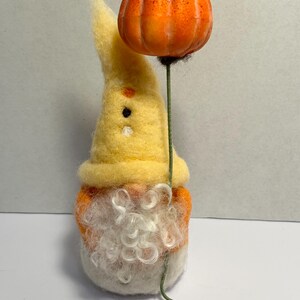 May include: A handmade felt gnome with a yellow hat and orange body, holding a small orange pumpkin on a green wire stem. The gnome has a white beard and a whimsical design, perfect for fall or Halloween decor.
