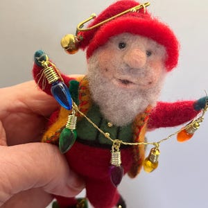 May include: A handmade felt Christmas ornament of a whimsical elf wearing a red hat and suit with a green and gold vest. The elf is holding a string of colorful Christmas lights. A gold safety pin and bell adorn the hat.
