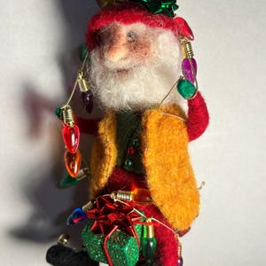 May include: A handmade Christmas ornament in the shape of a Santa Claus figure. The figure wears a red hat and coat, a yellow vest, and holds a string of colorful lights. A green glittery gift box is attached to the figure.