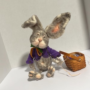 May include: A gray and white rabbit plush toy wearing a purple sweater and a green and orange bow. The rabbit is sitting with its ears up and is holding a small brown wicker basket.