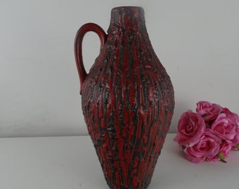 May include: A red and black ceramic vase with a textured surface and a handle. The vase is tall and narrow, with a rounded bottom and a flared top.