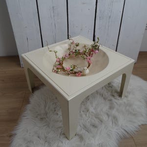 May include: A square, off-white coffee table with a recessed, circular top. The table has four straight legs. A decorative arrangement of pink flowers and greenery sits in the recessed area, along with a small white bird figurine. The table rests on a fluffy, white rug.