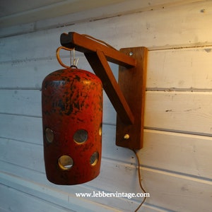 May include: A vintage wall lamp with a cylindrical red shade and circular cutouts. The lamp is supported by a wooden bracket and arm, mounted on a white wooden wall. The lamp has a copper wire handle. The website address is visible.