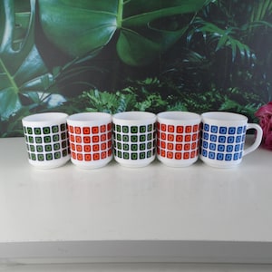 Könnte beinhalten: Satz von fünf Keramikbechern mit geometrischen Mustern. Die Becher sind weiß mit grünen, roten und blauen Quadraten.