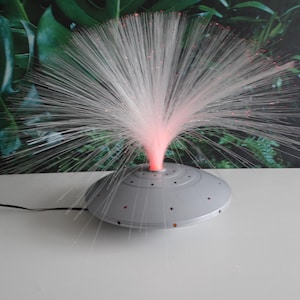 Op de afbeelding: Een glasvezellamp met een grijze basis en een witte, stralende lichtweergave. Het licht komt uit het midden, met rode en roze tinten. De lamp staat op een wit oppervlak tegen een achtergrond van groen gebladerte.