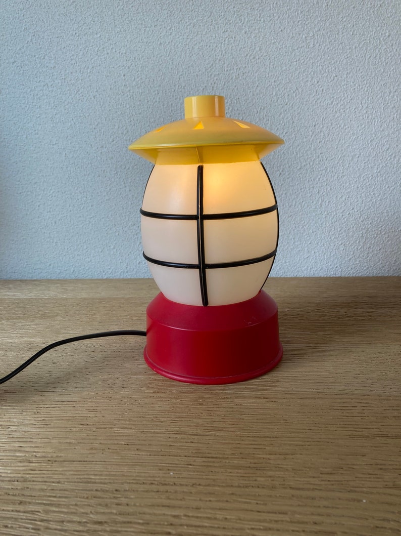 May include: A yellow and red plastic lamp shaped like a lantern with a white frosted glass shade and a black wire cage. The lamp is plugged in and lit.