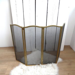 May include: A three-panel fireplace screen with a gold-coloured frame and a black mesh centre. The screen is standing on a light-coloured wooden floor and a white fluffy rug. The screen is designed to protect the area around a fireplace.