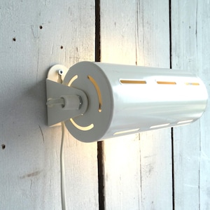 Può includere: Una lampada da parete cilindrica bianca con una serie di fessure orizzontali e ritagli curvi. La lampada è fissata a una staffa bianca e montata su una parete di legno chiaro. Un cavo elettrico bianco esce dalla lampada.