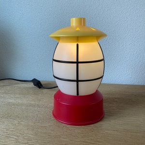 May include: A yellow and red plastic lamp shaped like a lantern with a white frosted glass globe and black metal bars. The lamp is plugged in and has a cord.