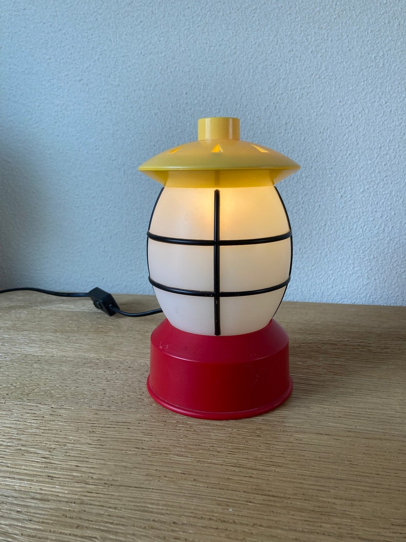 May include: A yellow and red plastic lamp shaped like a lantern with a white frosted glass globe and black metal bars. The lamp is plugged in and has a cord.
