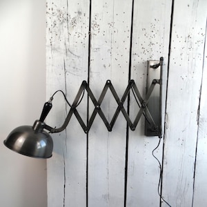 Könnte beinhalten: Industriell gestaltete Wandlampe mit Metalloberfläche. Die Lampe verfügt über einen verstellbaren Scherenarm und einen runden Schirm. Sie ist an einer weißen Holzoberfläche befestigt und hat ein schwarzes Kabel.
