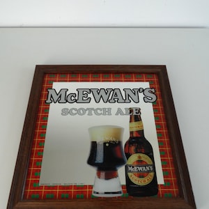 Op de afbeelding: Ingelijste reclame voor McEwan's Scotch Ale. De afbeelding toont een glas donker bier met schuim en een bierfles. De achtergrond heeft een rood en groen ruitpatroon. De lijst is bruin.