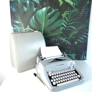 Puede incluir: Una máquina de escribir Hermes Media 3 gris vintage con teclas blancas y un maletín de transporte gris a juego. Un pequeño pincel descansa junto a la máquina de escribir. El fondo presenta una gran impresión de hojas tropicales verdes y negras.