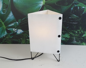 May include: A white, geometric table lamp with a textured, corrugated shade. The lamp has three black legs and three black fasteners on the side. A black power cord extends from the base. The background features a green leaf pattern.