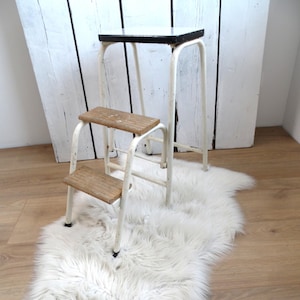 May include: A vintage step stool with a white metal frame and two wooden steps. The top step is a square with a black border. The stool is placed on a white faux fur rug. The background is a white wooden wall.