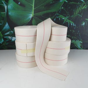 May include: Several rolls of off-white fabric tape with a red dotted line down the center. The rolls are stacked on top of each other, with some tape unrolled. The background features a green and black botanical print.