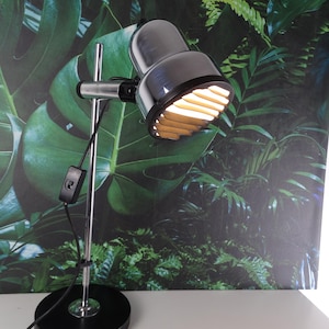 May include: A black and chrome desk lamp with a cylindrical head and a round base. The lamp has a silver arm and a black power cord. The lamp's head is angled downward, and the light is on. The background features a green and black tropical leaf pattern.