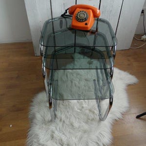 Puede incluir: Un teléfono naranja con dial rotatorio descansa sobre una mesa auxiliar de cristal de tres niveles con patas cromadas. La mesa se apoya sobre una alfombra de piel sintética blanca, creando una estética retro.