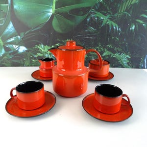 May include: A bright orange tea set featuring a central teapot, a lidded sugar bowl, a creamer, and two cups with matching saucers. Each piece has a smooth, glossy finish with a distinct black rim along the edges and a black interior lining.