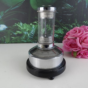 May include: A vintage barometer with a clear glass cylinder and a silver-colored metal base. The top of the cylinder reads "Thermometre" and "Hygrometre". The base has a black, round stand. The barometer measures atmospheric pressure.