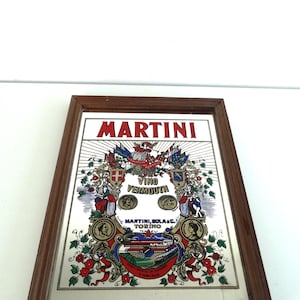 Op de afbeelding: Een ingelijste Martini-advertentie met de merknaam in het rood, en de woorden "Vino Vermouth". Het kunstwerk bevat een wapen, bloemelementen en een gebouwillustratie, alles in een bruine houten lijst.