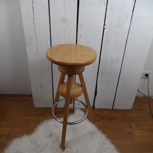 Pode incluir: Um banquinho de madeira de altura ajustável com um assento redondo e um apoio para os pés de metal. O banquinho é feito de madeira castanho claro e tem um design simples.