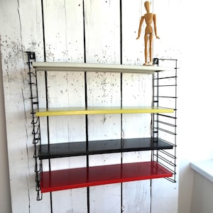 May include: A multi-coloured wall-mounted shelving unit with shelves in white, yellow, black, and red. The shelves are supported by a black wire frame. A wooden artist's mannequin stands on the top shelf. The unit is mounted on a white, weathered wooden panel.